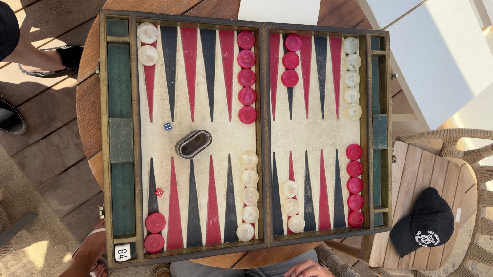 Un plateau de backgammon photographié d’en haut sur une table de terrasse, pions rouges et blancs, une casquette posée sur la chaise voisine.