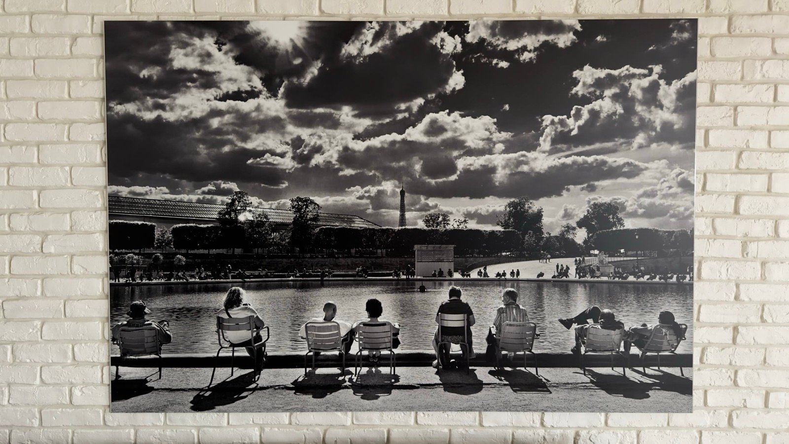 Photographie en noir et blanc accrochée sur un mur de briques blanches : des promeneurs assis de dos sur des chaises au bord d’un grand bassin, la tour Eiffel au loin sous un ciel chargé de nuages.